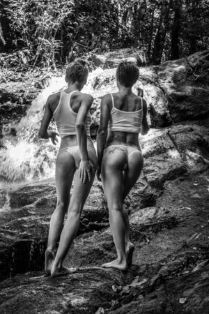 Back down view of two stunning girls in white tops and panties standing on the rock over beautiful waterfall background - black and white photoの写真素材