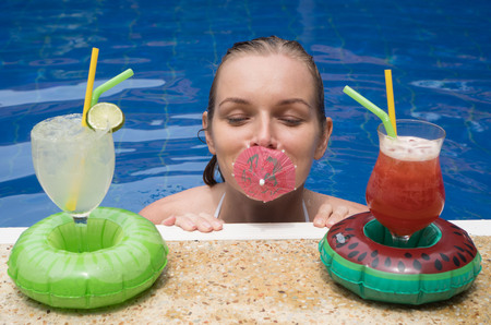 Happy smiling woman standing in the water with umbrella in her mouth and cocktails at the edge of...