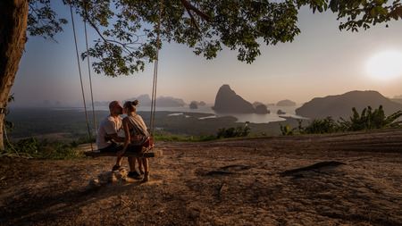 Kissing couple sitting on the swinging bench over beautiful tropical mountain landscape during sunriseの写真素材