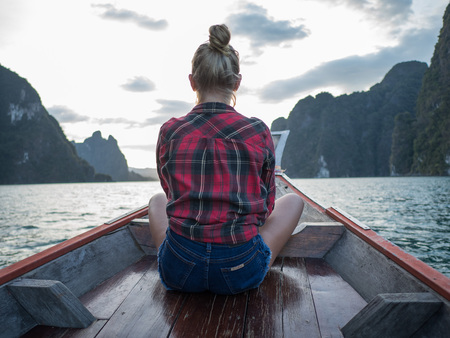 Back view of woman sitting in wood long tail boat over tropical limestone cliffs and sunset backgroundの写真素材