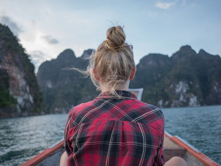 Back view of woman sitting in wood long tail boat over tropical limestone cliffs and sunset backgroundの写真素材