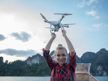 Woman sitting in wood long tail boat with raised hands holding drone over tropical limestone cliffs and sunset backgroundの写真素材