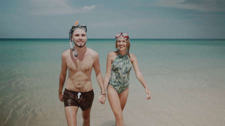 Young happy couple with snorkeling masks walking out from sea water, enjoying sunny summer day on...