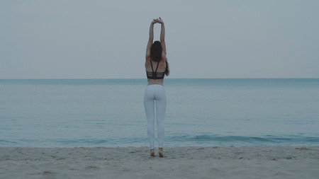 Back view of beautiful young girl exercising in the morning on the sandy beach over sea and sky...