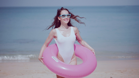 Beautiful young woman in swimsuit and sunglasses with pink inflatable ring having fun, enjoying sunny summer day on her beach tropical holidayの写真素材
