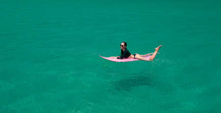 Aerial drone view of beautiful young woman in swimsuit and sunglasses with pink surfboard enjoying...