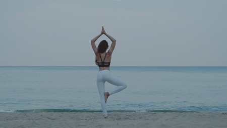 Back view of beautiful young girl exercising / doing yoga in the morning on the sandy beach over sea and sky backgroundの写真素材