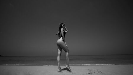 Beautiful young woman in swimsuit with snorkeling standing on the sandy beach over sea and sky background. Pretty girl enjoying sunny summer day on her beach tropical holiday black and white photoの写真素材