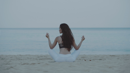 Back view of beautiful young girl exercising / doing yoga in the morning on the sandy beach over sea and sky backgroundの写真素材