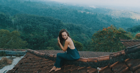 Aerial drone view of beautiful woman in long blue dress sitting on tiled red roof of the house...