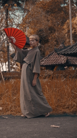 Beautiful fashion girl in kimono with red hand fan posing outdoor near traditional Balinese temple. Portrait of stylish hipster female outdoor - vertical photo imageの写真素材
