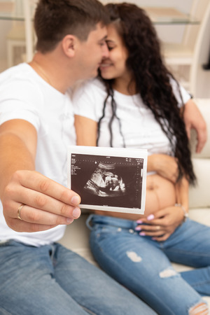 Portrait of cheerful married and pregnant couple holding ultrasound scan while sitting on sofa at...