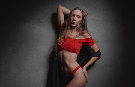 Beautiful young woman in red bikini and black overall posing over gray wall background
