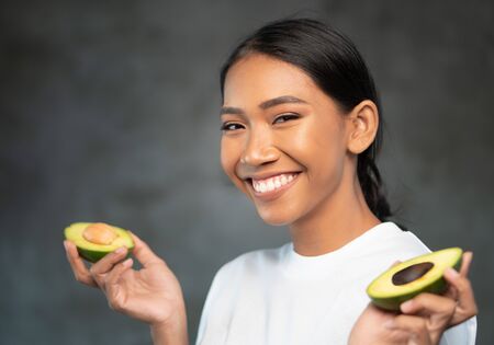 Portrait of beautiful young smiling woman in white t-shirt holding halves of avocado over concrete...
