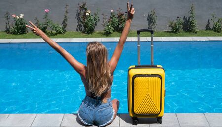 Back view of happy woman with yellow luggage sitting by the swimming pool. Summer, holiday, travel...