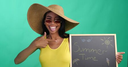 Bright summer portrait of beautiful smiling woman in yellow swimsuit, hat and red sunglasses holding chalk board with Summer time writing isolated over green screen backgroundの写真素材