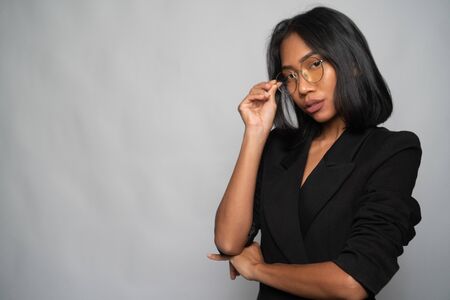 Portrait of attractive young Asian woman in elegant black blazer and glasses posing over light gray wall background. Mock up copy spaceの写真素材