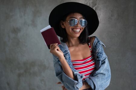 Portrait of smiling young Asian woman holding passport isolated over concrete wall background. People lifestyle, travel conceptの写真素材