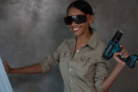 Portrait of attractive young girl builder with drill in overall and protective glasses standing on...