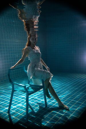 Beautiful woman with long red hair posing underwater on the chair in white dressの写真素材