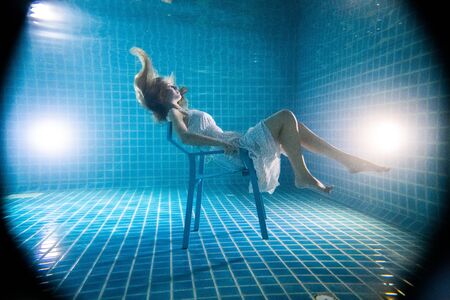Beautiful woman with long red hair posing underwater on the chair in white dressの写真素材