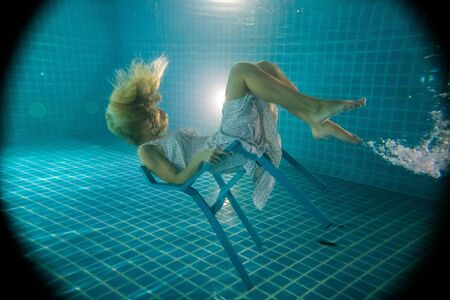 Beautiful woman with long red hair posing underwater on the chair in white dressの写真素材