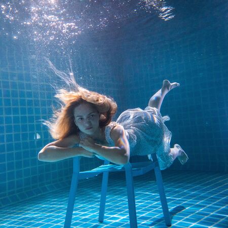 Beautiful woman with long red hair posing underwater on the chair in white dressの写真素材
