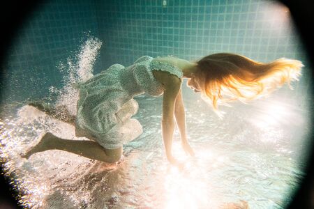 Beautiful woman with long red hair posing underwater in white dressの写真素材