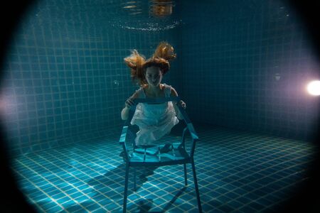 Beautiful woman with long red hair posing underwater on the chair in white dressの写真素材