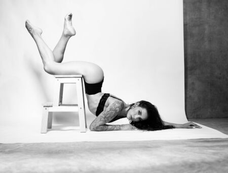 Beautiful acrobatic in black lingerie posing on the wooden chair stepladder in the studio over white wall background - black and white photographyの写真素材