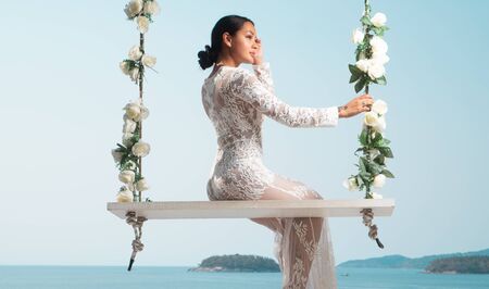Beautiful woman in white lace dress posing on the outdoor swing with flowwers over sea and sky backgroundの写真素材
