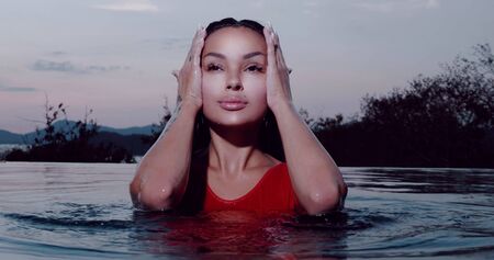 Portrait of beautiful brunette woman in red swimsuit posing in outdoor pool over sunset sky backgroundの写真素材