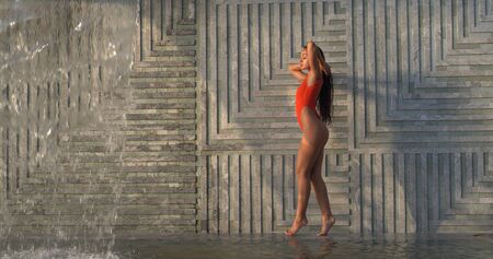 Beautiful brunette woman in red swimsuit posing near outdoor pool over wall with waterfall backgroundの写真素材