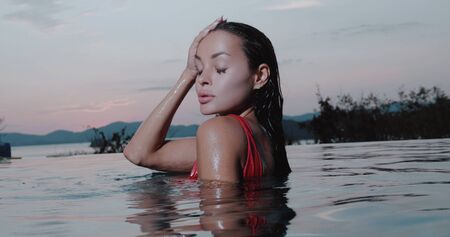 Portrait of beautiful brunette woman in red swimsuit posing in outdoor pool over sunset sky backgroundの写真素材