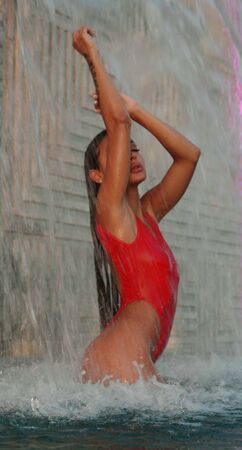Beautiful brunette woman in red swimsuit posing in outdoor pool over wall with waterfall backgroundの写真素材