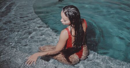 Beautiful brunette woman in red swimsuit posing in outdoor pool in the eveningの写真素材