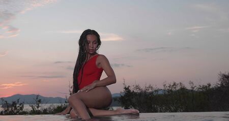 Beautiful brunette woman in red swimsuit posing by outdoor pool over sunset sky backgroundの写真素材