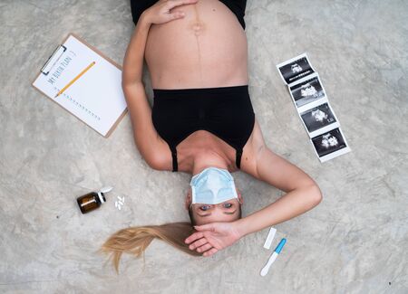 Pregnant woman in protective mask against flu and viruses on grey wall background lying scared on the floor. Healthcare conceptの写真素材