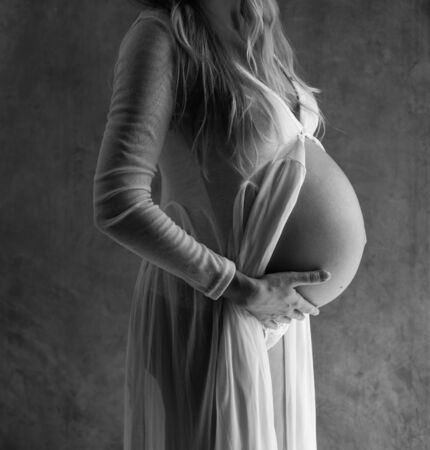 Closeup pregnant woman touching her belly while standing over gray wall background. Maternity...