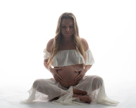 Pretty pregnant woman touching her belly while sitting over white background. Maternity conceptの写真素材