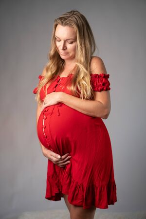 Pretty pregnant woman in red dress holding her belly while standing over gray wall background. Maternity conceptの写真素材