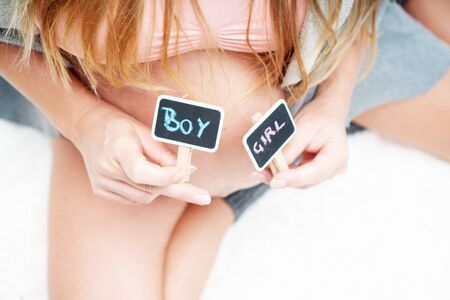 Cropped image of pregnant woman holding mini blackboards with written boy and girl while sitting on her bed at home. Maternity, pregnancy, new life and family planning conceptの写真素材