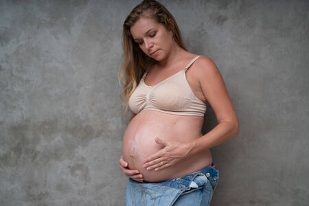 Pretty pregnant woman in denim jumpsuit and bra applying lotion on her belly. Maternity, pregnancy, new life and health care conceptの写真素材