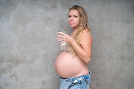 Pretty pregnant woman in denim jumpsuit and bra holding glass of water while standing over gray wall background. Maternity, pregnancy, new life healthy food conceptの写真素材