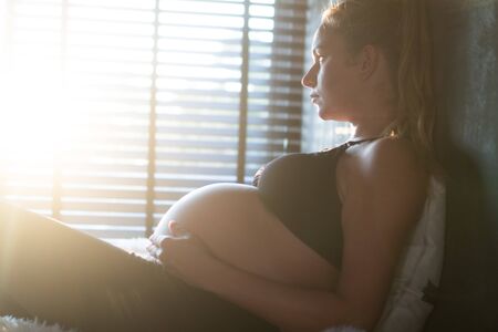 Pretty pregnant woman in black leggins and bra enjoying sunlight while relaxing on her bed at home. Maternity conceptの写真素材
