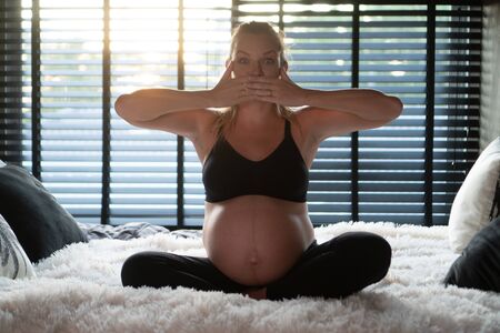 Pretty pregnant woman in black leggins and bra covering mouth with hands while sitting on her bed at home. Maternity, health care, expression of fear, secret conceptの写真素材