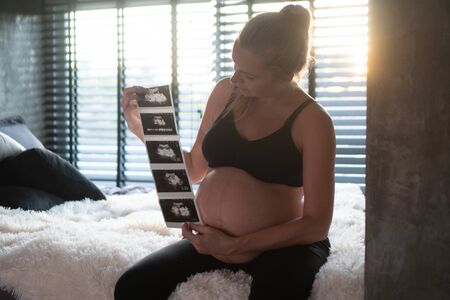 Pretty pregnant woman holding ultrasound photos while sitting on bed and relaxing at home. Maternity, pregnancy, new life and family planning conceptの写真素材