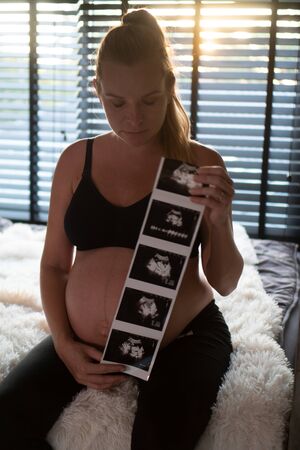 Pretty pregnant woman holding ultrasound photos while sitting on bed and relaxing at home. Maternity, pregnancy, new life and family planning conceptの写真素材