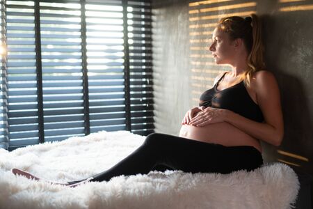 Pretty pregnant woman in black leggins and bra enjoying sunlight while relaxing on her bed at home. Maternity conceptの写真素材