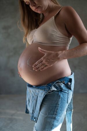 Pretty pregnant woman in denim jumpsuit and bra applying lotion on her belly. Maternity, pregnancy, new life and health care conceptの写真素材
